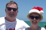 A man and woman in white t-shirts and a santa hat hug and smile on a beach.