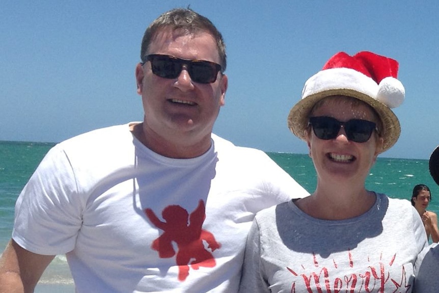 A man and woman in white t-shirts and a santa hat hug and smile on a beach.