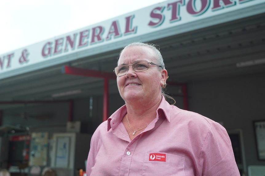 A woman in a pink collared shirt with an Australia post logo,  standing outside a small general store.