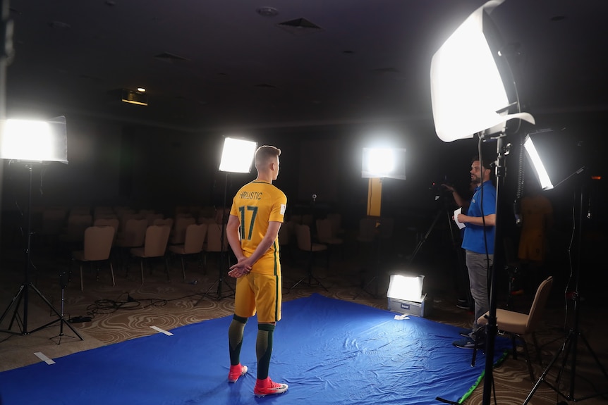 A male soccer player wearing yellow and green stands in a room surrounded by lights