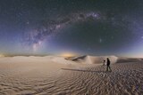 A photographer waits by her tripod with the camera pointed to the sky beneath the Milky Way in front of sand dunes in the desert