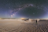 A photographer waits by her tripod with the camera pointed to the sky beneath the Milky Way in front of sand dunes in the desert