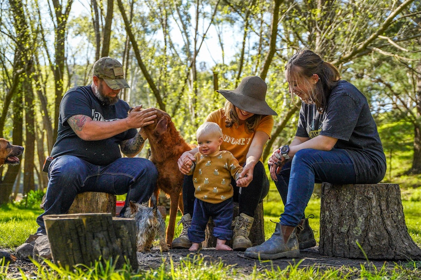 A young family sit on wooden logs surrounded by leafy trees.