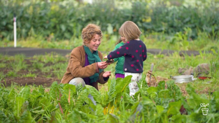 Woman kneeling in vegie garden with two small children passing her leaves