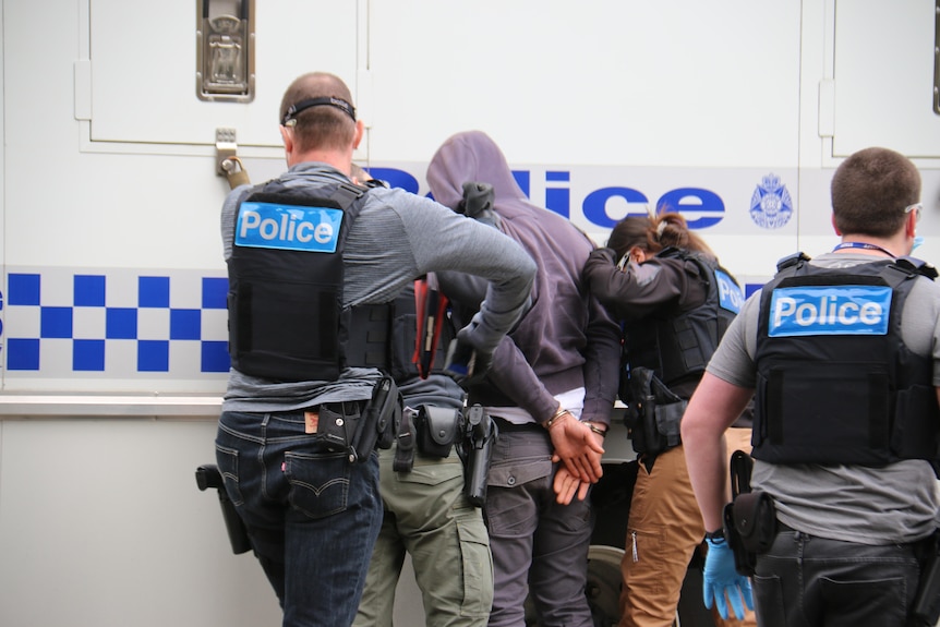 Police handcuff a man in a hooded jumper as they stand beside a marked police vehicle.