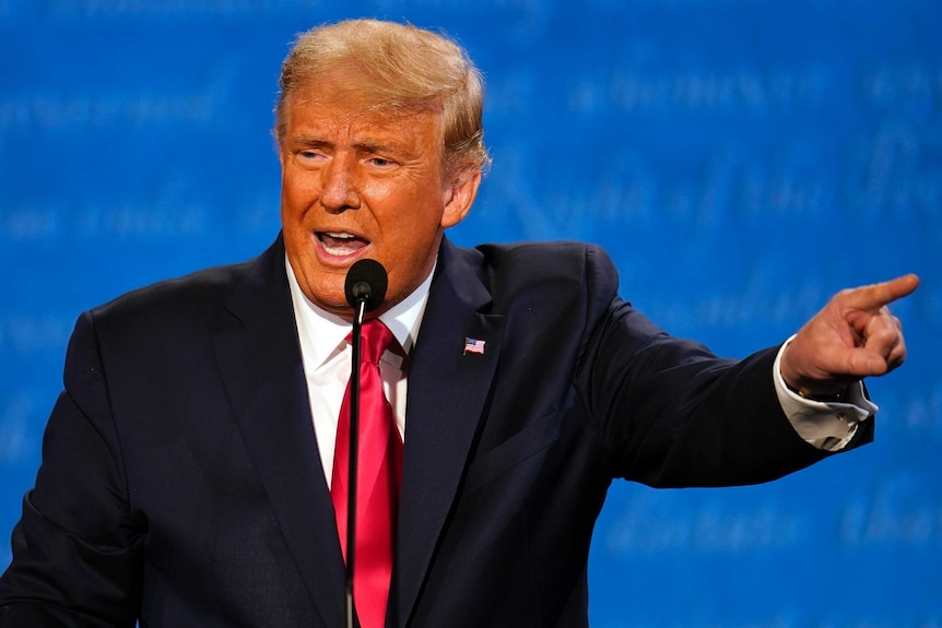 A man with blonde hair and wearing a dark suit with a red tie and United States flag pin points his finger