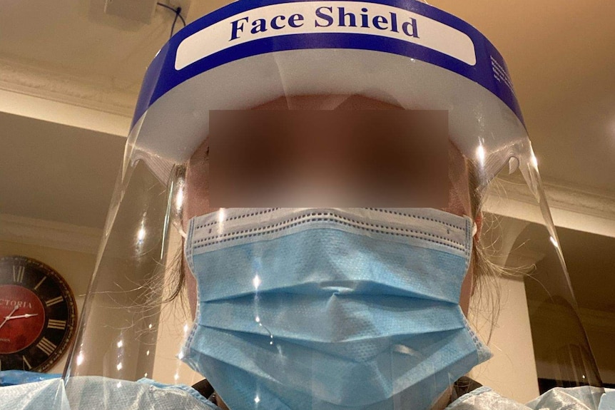 A woman wears a surgical mask, face shield and gown with her eyes blurred out.