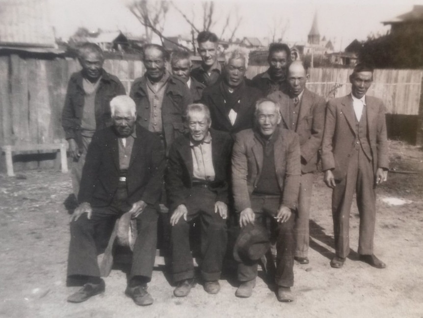 Group of 11 men in a yard with a wooden fence and rooftops in the background.