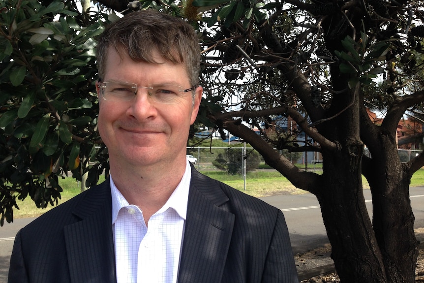 A smiling, bespectacled man in a black blazer and white shirt, standing in front of a tree.