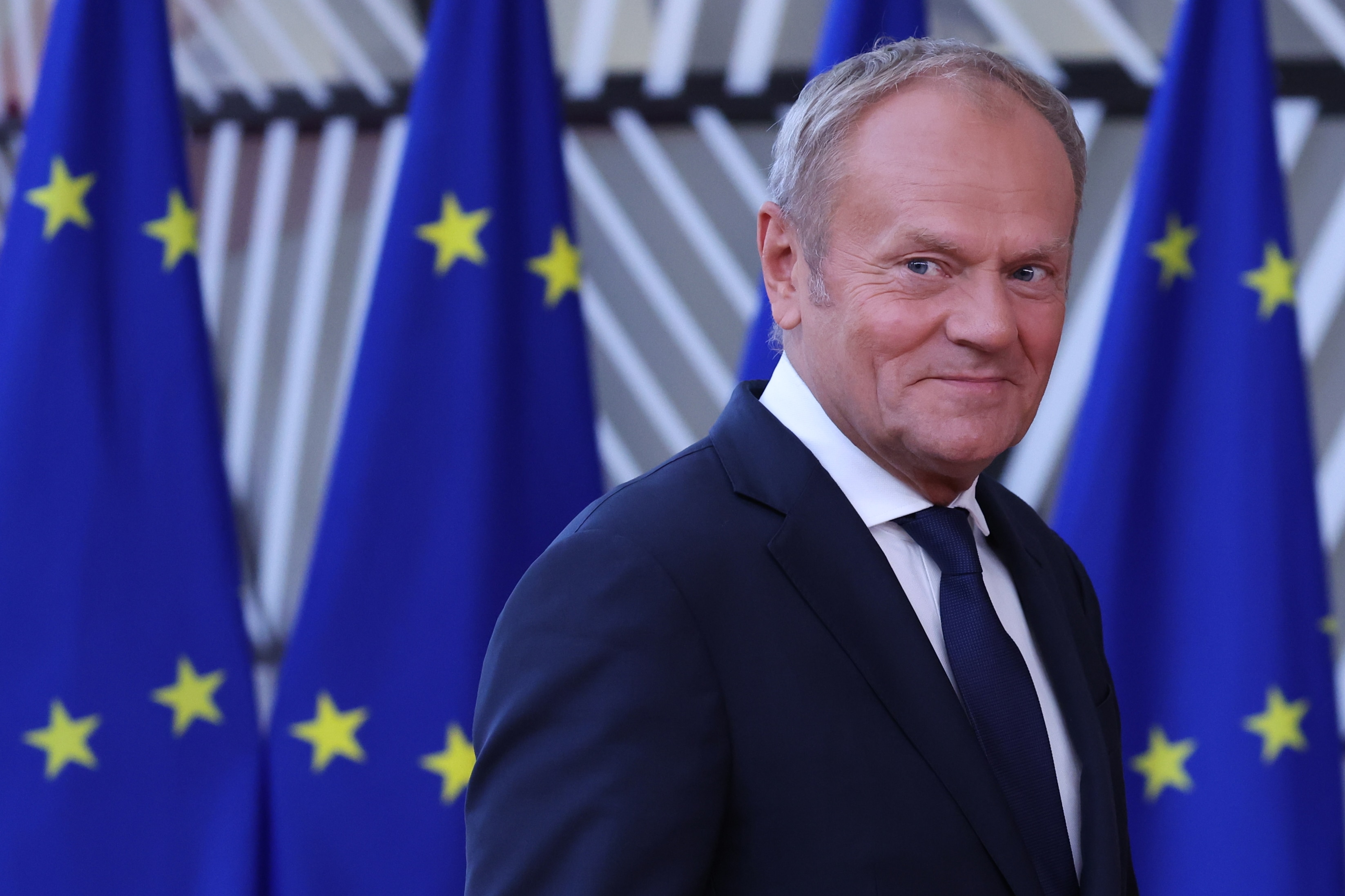 Donald Tusk in a black suit and tie, seen on a blurred background.