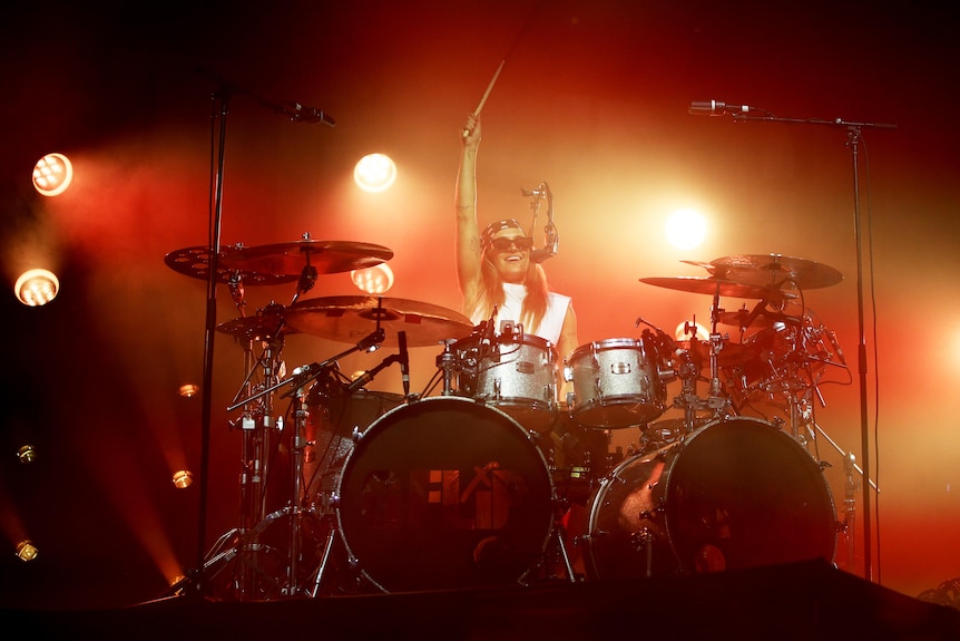 G Flip behind a drum kit on stage with their right arm up in the air, singing into a microphone. Red lights are behind them