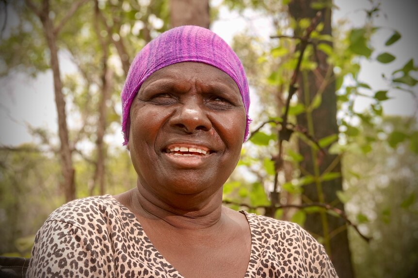 An Aboriginal women in a bright purple bandana, standing in front of some mangroves. 