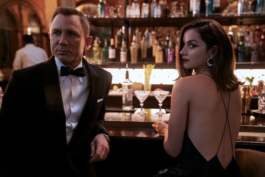 Man with cropped blonde hair wears black tuxedo beside brunette woman with dark lipstick and low-backed dress at a bar
