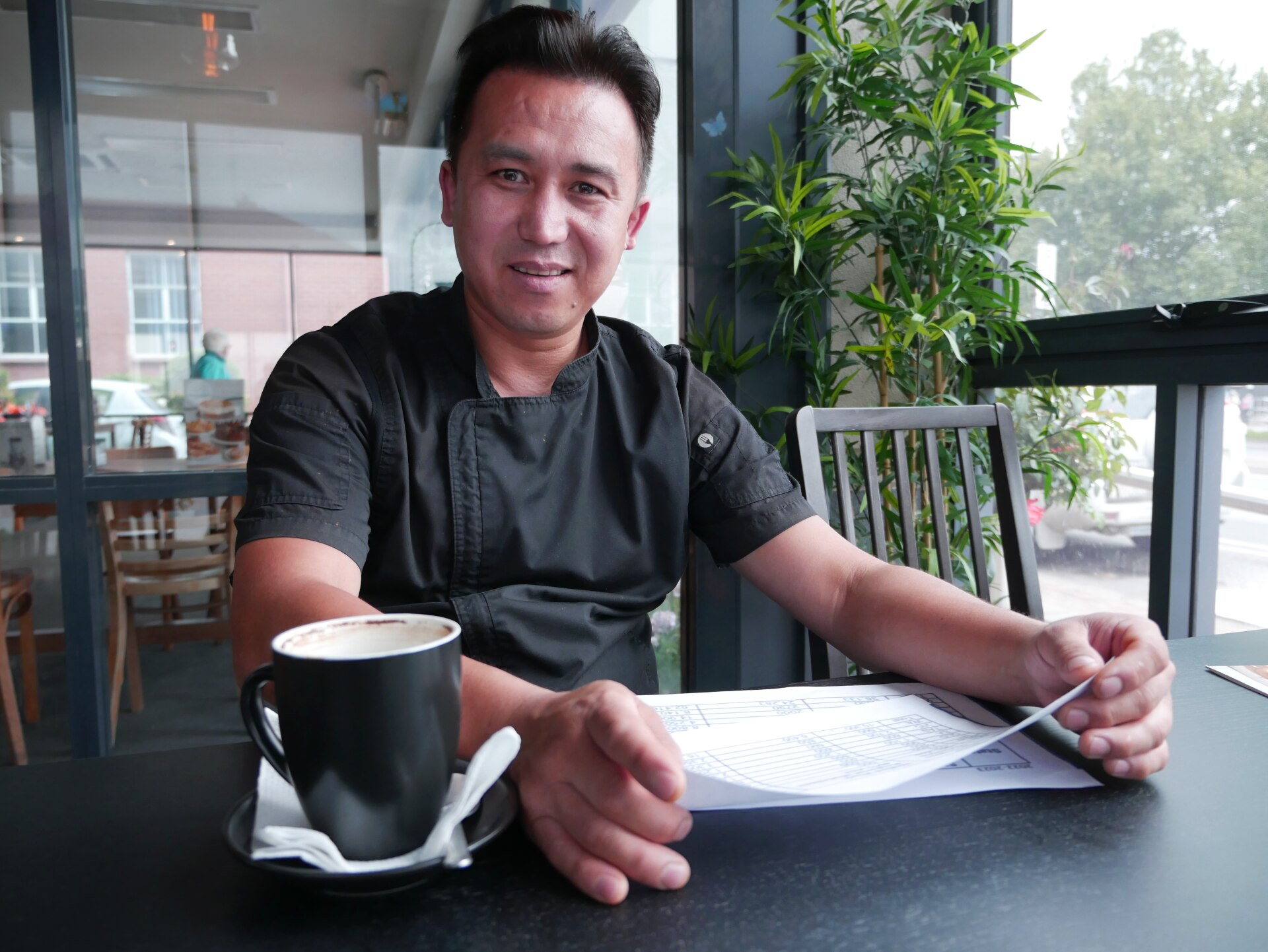 A Hazara man dressed in commercial cookery black sits at a table with an empty coffee mug at hand. He's looking at the camera.  