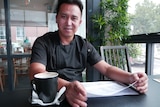 A Hazara man dressed in commercial cookery black sits at a table with an empty coffee mug at hand. He's looking at the camera.  