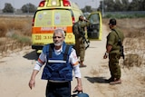 An older middle-aged man in a blue bulletproof vests walks in the desert near a bright yellow ambulance and two soldiers.