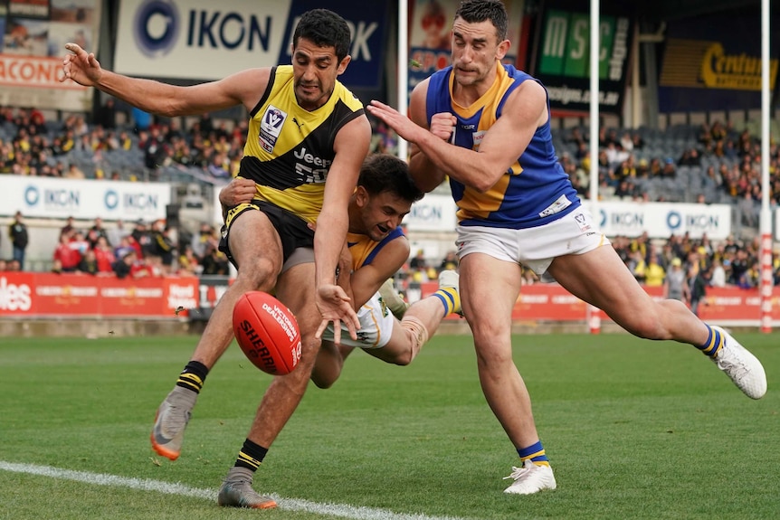 Marlion Pickett kicks the ball under pressure from two players in blue and yellow singlets