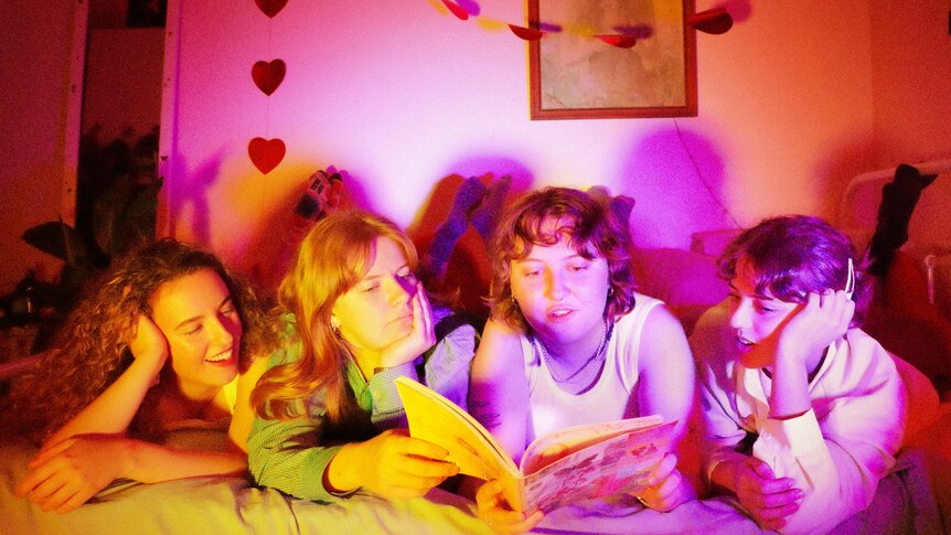 Four people lie on their stomachs on a bed looking at a magazine in a bedroom lit pink and yellow