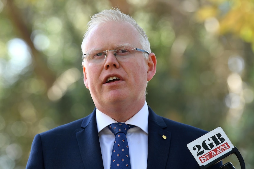 A light-haired, bespectacled politician in a dark suit stands outside speaking to the media.