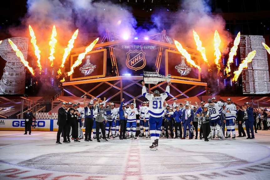 The Tampa Bay Lightning team celebrate winning the Stanley Cup on the ice rink with pyrotechnics happening behind.