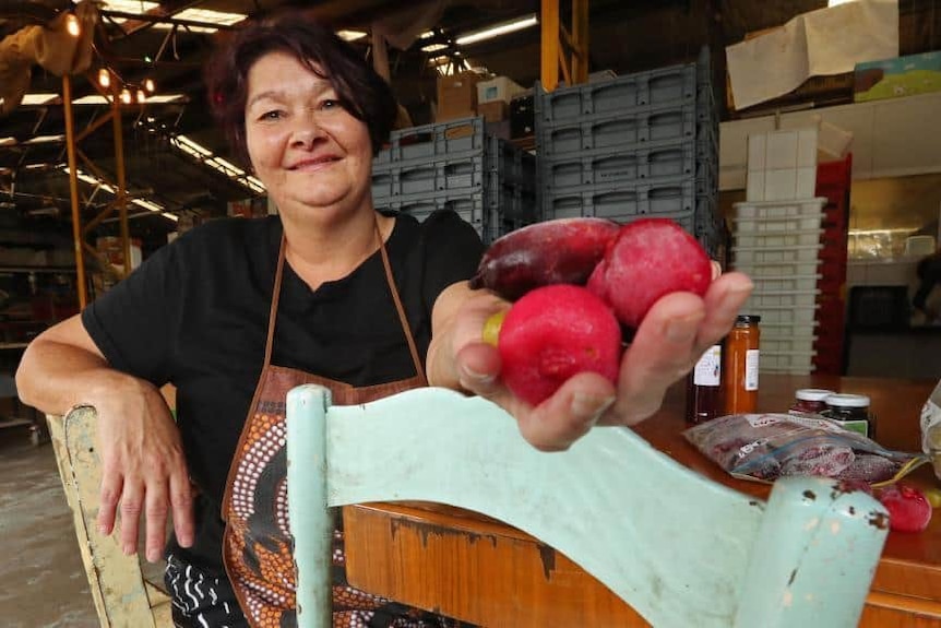 Aunty Dale Chapman, with short brown hair, smiling widely, holds hand out in which several large pink fruits sit.
