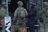 Heavily armed police in combat gear stand near an armed vehicle, with a police dog.