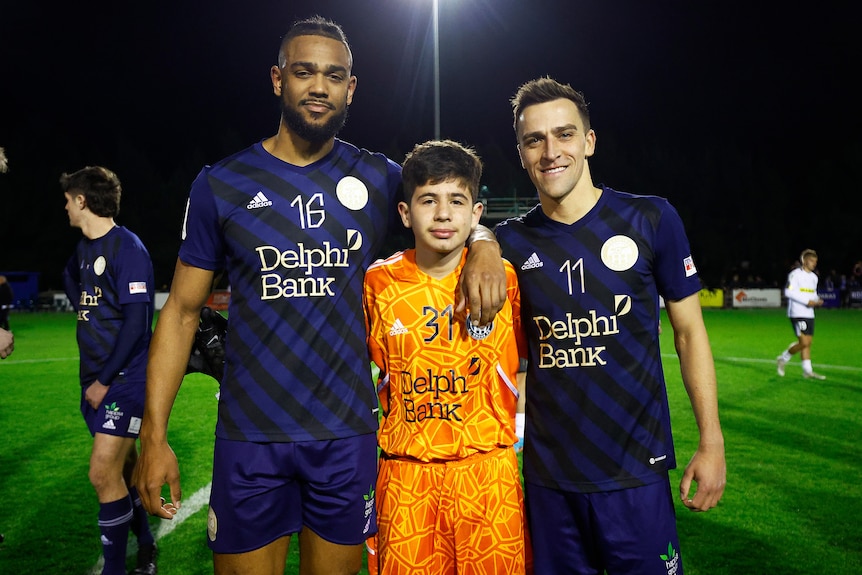 A boy in an orange goalkeeper's kit stands between two adult football players after an Australia Cup game.