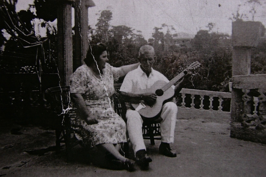 An old vintage photo of an old man plays guitar while an old woman puts her arm around him