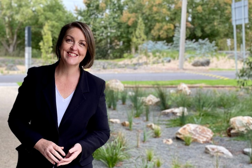 A smiling, dark-haired woman wearing a dark blazer stands in a bushy area.