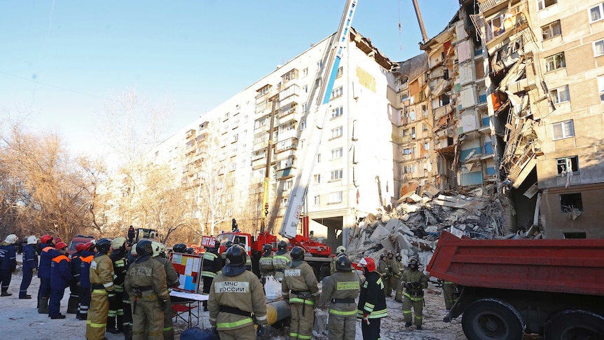 On a clear sky, a crane lifts concrete debris from an apartment block which has partially collapsed with firefighters looking on