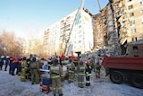 On a clear sky, a crane lifts concrete debris from an apartment block which has partially collapsed with firefighters looking on
