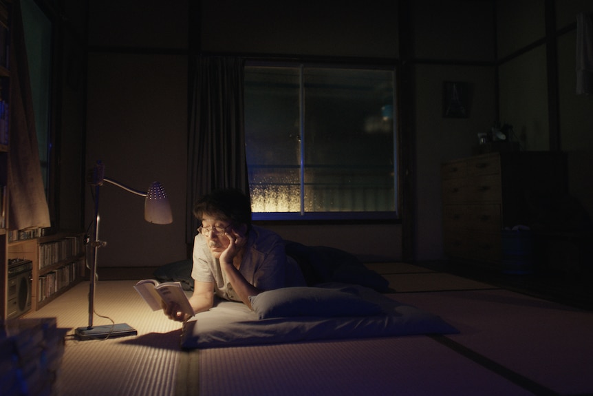 A film still of Kōji Yakusho, a 68-year-old Japanese man, lying down reading a book by lamplight, his chin in his hand.