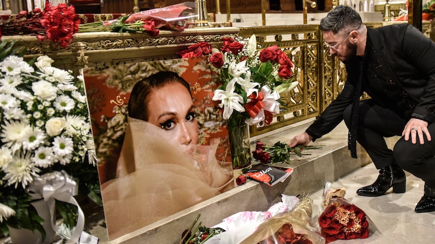 A person bends down to leave flowers next to an image of a woman surrounded by other flowers at the front of a church