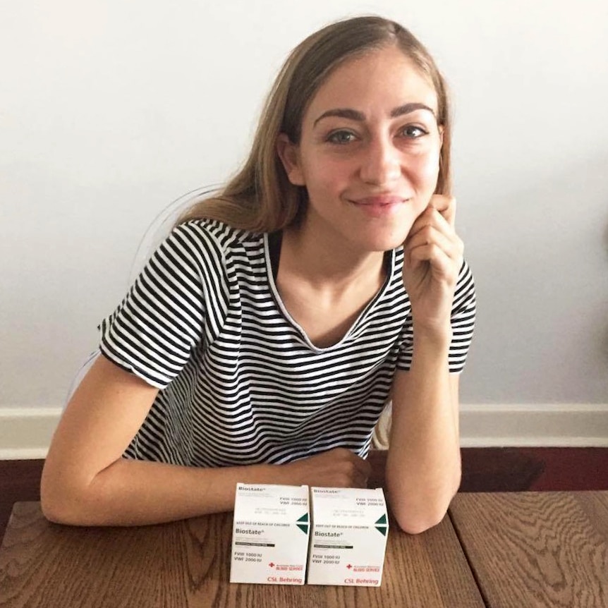 Chloe Christos sits smiling with two boxes of blood medication on the table in front of her.