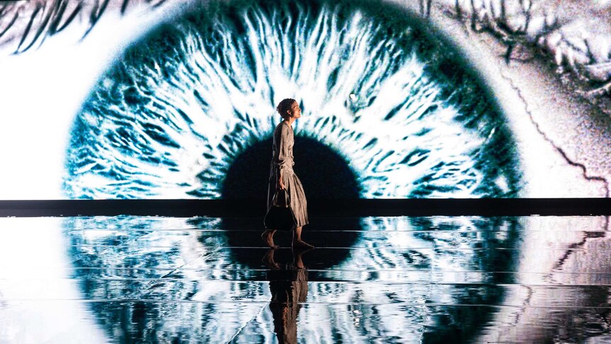 A stage with a woman in profile walking across it, a projection of an enormous eye in the background