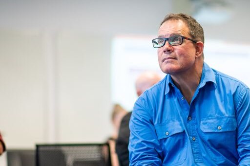 A man in a collared blue work shirt and glasses sitting and watching something off-camera.