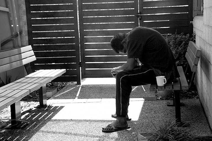 A black and white photo of a man sitting slumped on a bench with a coffee cup beside him.