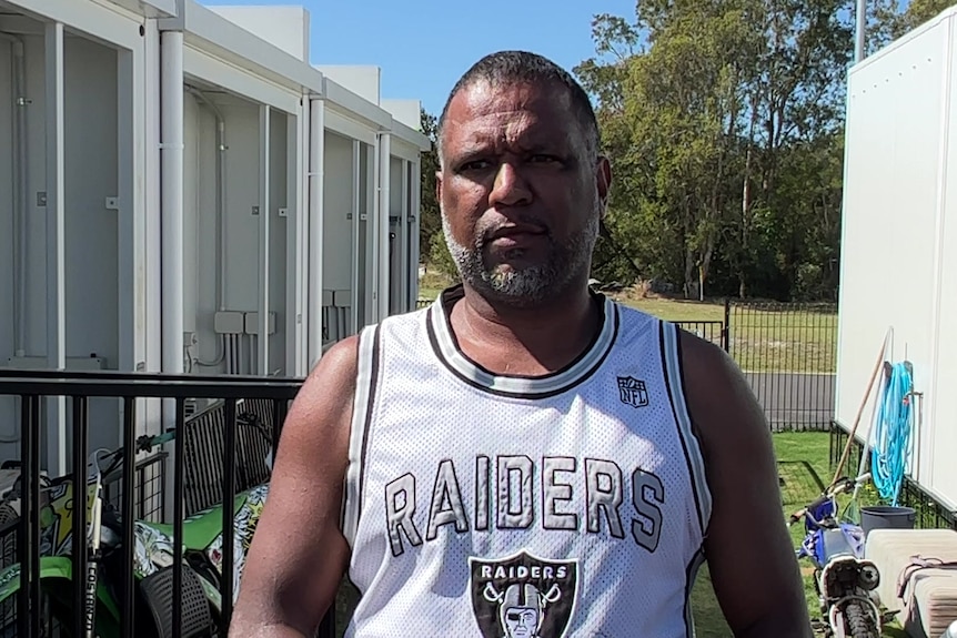 An Indigenous man wearing a tank top, standing between two buildings.