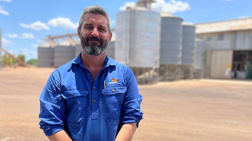 Man with blue shirt and grey beard standing in front of grey sheds and silos smiling