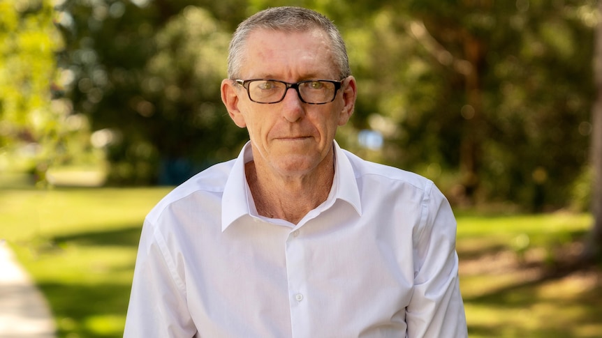 A middle-aged white man with short hair, a white shirt and glasses standing in a park