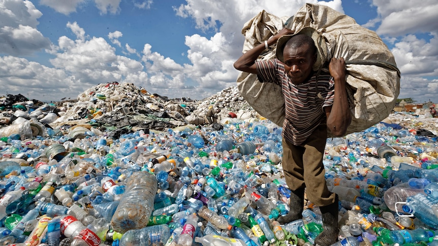 A man walks on a mountain of plastic bottles as he carries a sack of them.