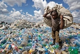 A man walks on a mountain of plastic bottles as he carries a sack of them.