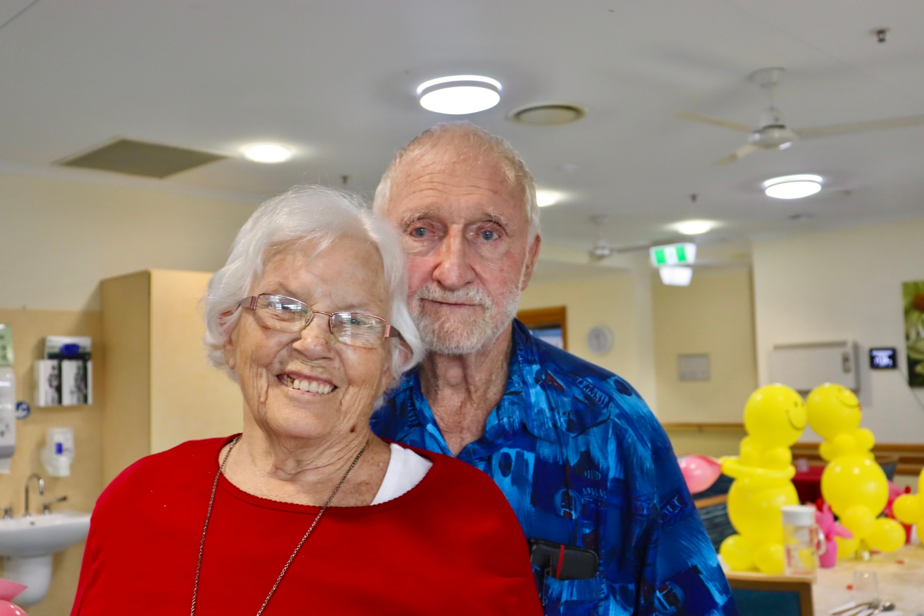 An elderly woman and man in a nursing home room with yellow balloons in the background