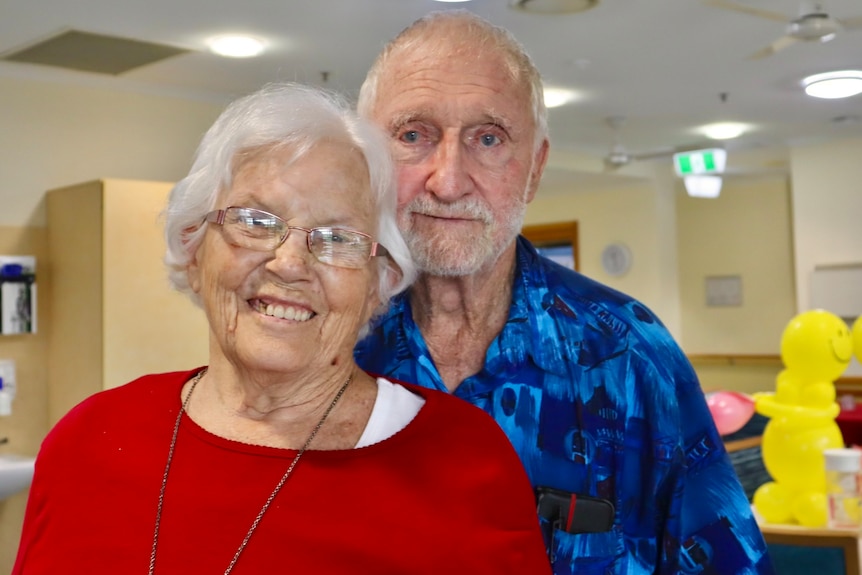 An elderly woman and man in a nursing home room with yellow balloons in the background