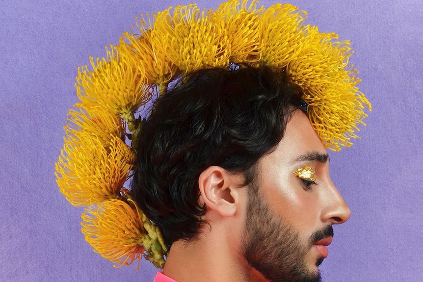 A man stands side on with yellow flowers shaped into a mohawk with gold eye shadow.
