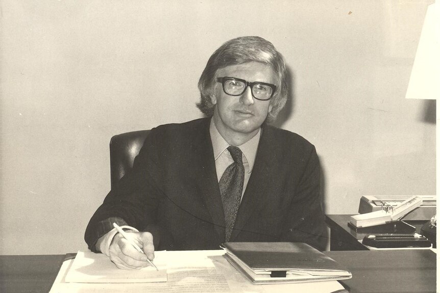 A black and white photo take in the 1970s of a young man wearing a suit sitting at a desk.