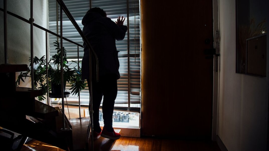 A woman stands inside a house, looking through the blinds.