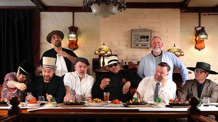 The eight members of Fat Freddy's Drop sit around a table sharing a meal