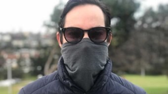 A man wears a turtleneck pulled up over his mouth and nose as a facemask, under sunglasses.