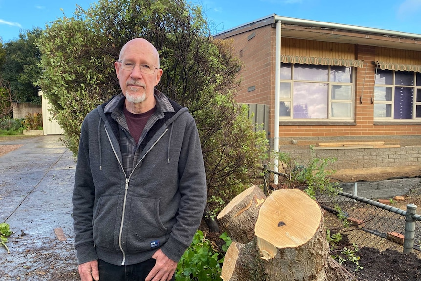 A man stands next to a tree stump in front of his home.